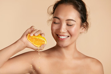 Wall Mural - Beauty. Woman with radiant face skin squeezing orange in hand portrait. Beautiful smiling asian girl model with natural makeup, glowing facial skin and citrus fruit. Vitamin C cosmetics concept
