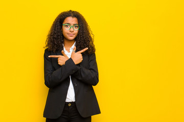 black business woman looking puzzled and confused, insecure and pointing in opposite directions with doubts against orange wall