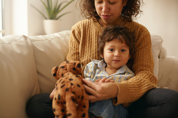 Wall Mural - Indoor shot of happy caring young Latin woman sitting on sofa with adorable toddler on her lap playing with stuffed animal spending nice time together. Mother and son bonding in living room