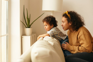 Casually dressed young Latin female with curly hair sitting on couch with her two year son looking through window, watching life outside, having curious facial expression, studying world around him