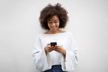 Wall Mural - smiling young african american woman looking at cellphone by white wall