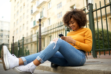 Wall Mural - cool young african american woman sitting outside on street with mobile phone