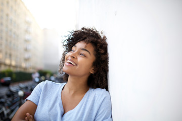 Wall Mural - smiling young african american woman leaning against wall outside and looking away