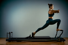 Young Girl Doing Pilates Exercises With A Reformer Bed. Beautiful Slim Fitness Trainer On A Reformer Gray Background, Low Key, Art Light, Copy Space Advertising Banner