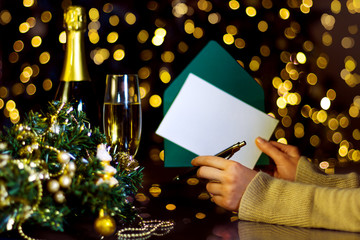 Wall Mural - Female hands are holding envelope with blank sheet of paper for greeting card. Decorated wreath, glass with champagne on table. Garland with yellow light bulbs are shining. New year, christmas mood.