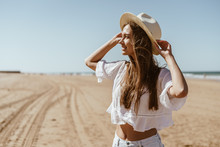 Attractive European Woman Stands On The Beach And Looks Into The Distance, Holding Her Hat With Her Hands
