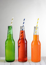 Three Bottles Of Soda With Straws On A Wood Table, Lemon-Lime, Strawberry And Orange Flavors.