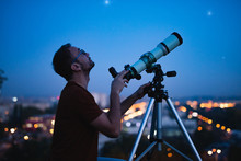 Astronomer With A Telescope Watching At The Stars And Moon With Blurred City Lights In The Background.