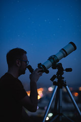 Wall Mural - astronomer with a telescope watching at the stars and moon with blurred city lights in the backgroun