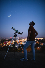 Wall Mural - astronomer with a telescope watching at the stars and moon with blurred city lights in the backgroun
