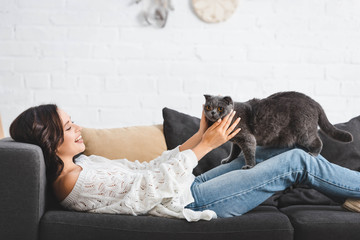 Wall Mural - brunette woman lying on sofa with scottish fold cat in cozy living room