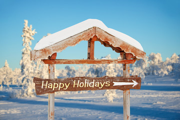 Wall Mural - Happy holidays written on a wooden direction sign, blue sky and winter snowy tree landscape background seasons greetings christmas card