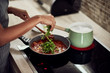 © nenadaksic - Mixed race woman in apron standing next to stove, stirring tomato sauce and adding rocket. On stove is pot with boiling pasta.