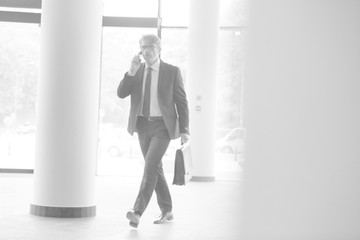 Wall Mural - Black and white photo of Mature businessman walking while talking on smart phone in the lobby