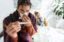 Sick Man With Fever Holding Thermometer And Napkin In Bedroom With Woman Behind