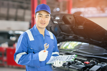 car service, repair, maintenance  concept - auto mechanic man or Smith writing to the clipboard at warehouse workshop, technician doing the checklist for repairs engine a car in the garage with laptop