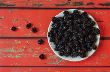 Ripe blackberries in a white plate on a red wooden background close-up. Fresh black berries still life.