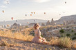 © biotin - woman is watching on scenery view with rising balloons on sunrise. Girl in gorgeous pink long dress sit on hill looking at large number of air balls. Fabulous Cappadocia mountains landscapes Turkey