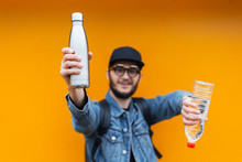 Portrait Of Cheerful Young Hipster, Holding Steel Thermo Eco Bottle For Water And Plastic Bottle, On Yellow Or Orange Background. Say No To Plastic.