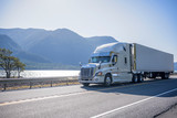 Big rig semi truck with chrome accessories transporting frozen cargo in refrigerated semi trailer moving on the road along river with bewitching view on the opposite bank