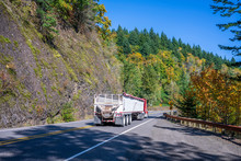 Red Big Rig Semi Truck With Bulk Semi Trailer Running Downhill On The Winding Autumn Road With Rock Mountain Wall And Forest