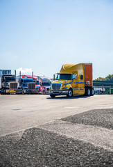 Yellow big rig semi truck with orange semi trailer driving on the truck stop driving out of a line of parked semi trucks