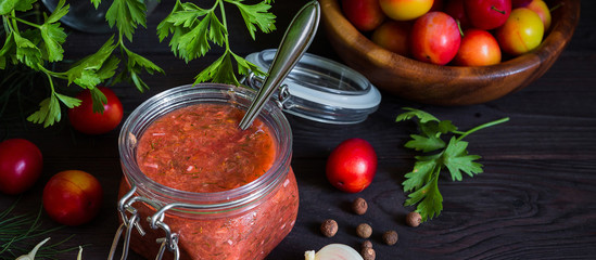 Tkemali sauce from red plums and ingredients for cooking garlic, parsley, dill, pepper with copy space on a dark wooden background