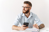 Fototapeta Na ścianę - Image of happy young man holding smartphone while working in office