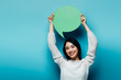 © LIGHTFIELD STUDIOS - smiling brunette asian woman holding green speech bubble on blue background