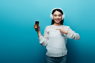 Wall Mural - smiling asian woman with headphones pointing with finger at smartphone with business app on blue background