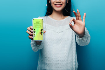 Wall Mural - cropped view of smiling asian woman in white blouse showing ok sign and smartphone with best shopping app on blue background