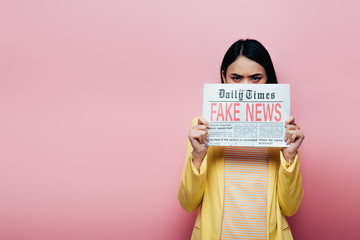 Wall Mural - sad asian woman in yellow outfit holding newspaper with fake news isolated on pink