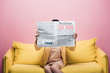 © LIGHTFIELD STUDIOS - asian woman holding newspaper with fake news in front of face while sitting on yellow sofa isolated on pink