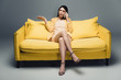displeased asian businesswoman sitting on yellow sofa with crossed legs and talking on smartphone on grey background