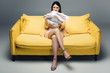 © LIGHTFIELD STUDIOS - aggressive asian businesswoman sitting on yellow sofa and reading newspaper with fake news on grey background