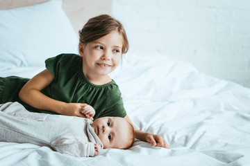 Wall Mural - smiling child holding hand of little sister lying on white bedding