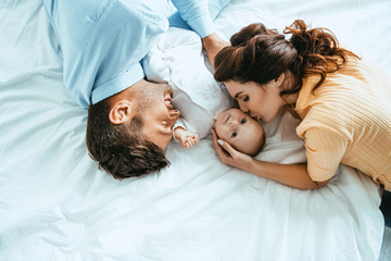 Wall Mural - Happy mother kissing baby while lying on bed near smiling husband