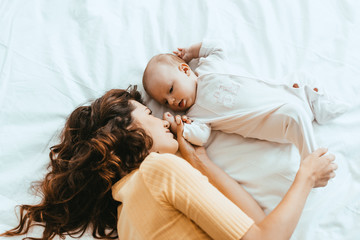 Wall Mural - top view of mother touching leg of cute little daughter while lying on white bedding