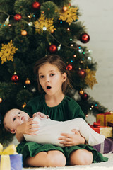 Wall Mural - amused child looking at camera while sitting near christmas tree and holding cute little sister