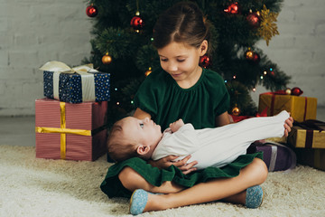 Wall Mural - smiling kid holding cute infant while sitting near Christmas tree and gift boxes