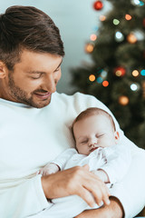 Wall Mural - happy father holding sleeping baby near christmas tree