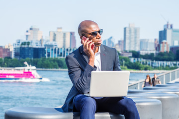 Wall Mural - Young African American Businessman traveling, working in New York, wearing blue blazer, sunglasses, sitting by East River, working on laptop computer, talking on cell phone. Brooklyn on far background