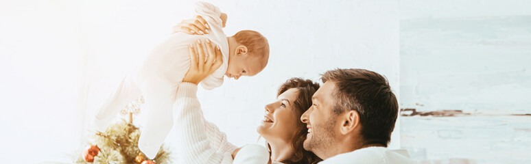 Wall Mural - panoramic shot of smiling man standing near wife holding cute infant above head