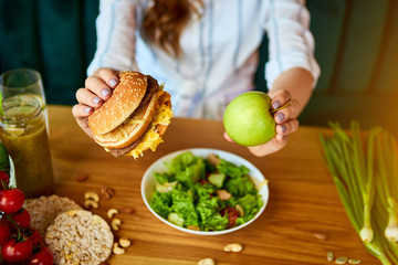 Wall Mural - Beautiful young woman decides eating hamburger or apple in kitchen. Cheap junk food vs healthy diet