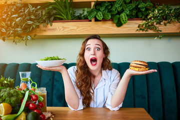 Wall Mural - Beautiful young woman decides eating hamburger or fresh salad in kitchen. Cheap junk food vs healthy diet