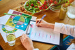 © bondvit - Woman dietitian in medical uniform with tape measure working on a diet plan sitting with different healthy food ingredients in the green office on background. Weight loss and right nutrition concept
