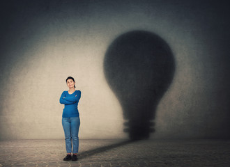 Confident woman, keeps arms crossed, casting a lightbulb shape shadow on the wall. Ambition and business idea concept. Genius innovation, motivation and inner power symbol.