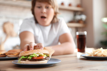 Wall Mural - Emotional overweight boy reaching for burger at table in kitchen