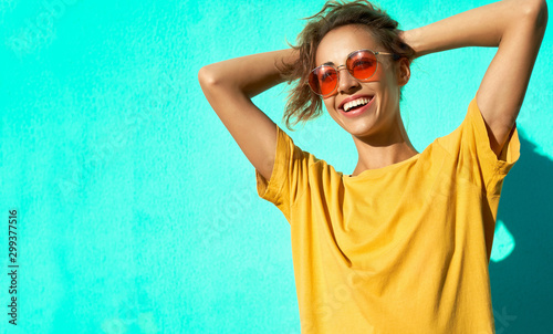 Cuadros en Lienzo Fashionable cheerful young woman posing on blue background, wearing yellow t-short and trendy red eyeglasses