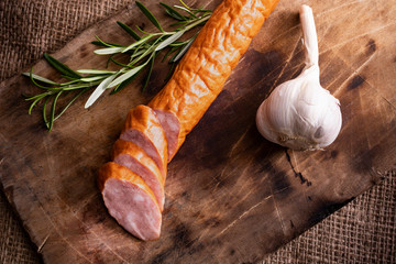 Smoked pork sausage on wood cutting board and jute fabric. A composition with appetizing sausage, garlic and rosemary.